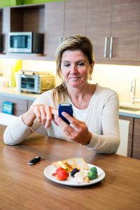 A diabetes patient checks her blood sugar levels on a glaucometer. The woman is sitting a home or at work in a modern kitchen or cafeteria setting. A plate of healthy food is arranged in front of her.